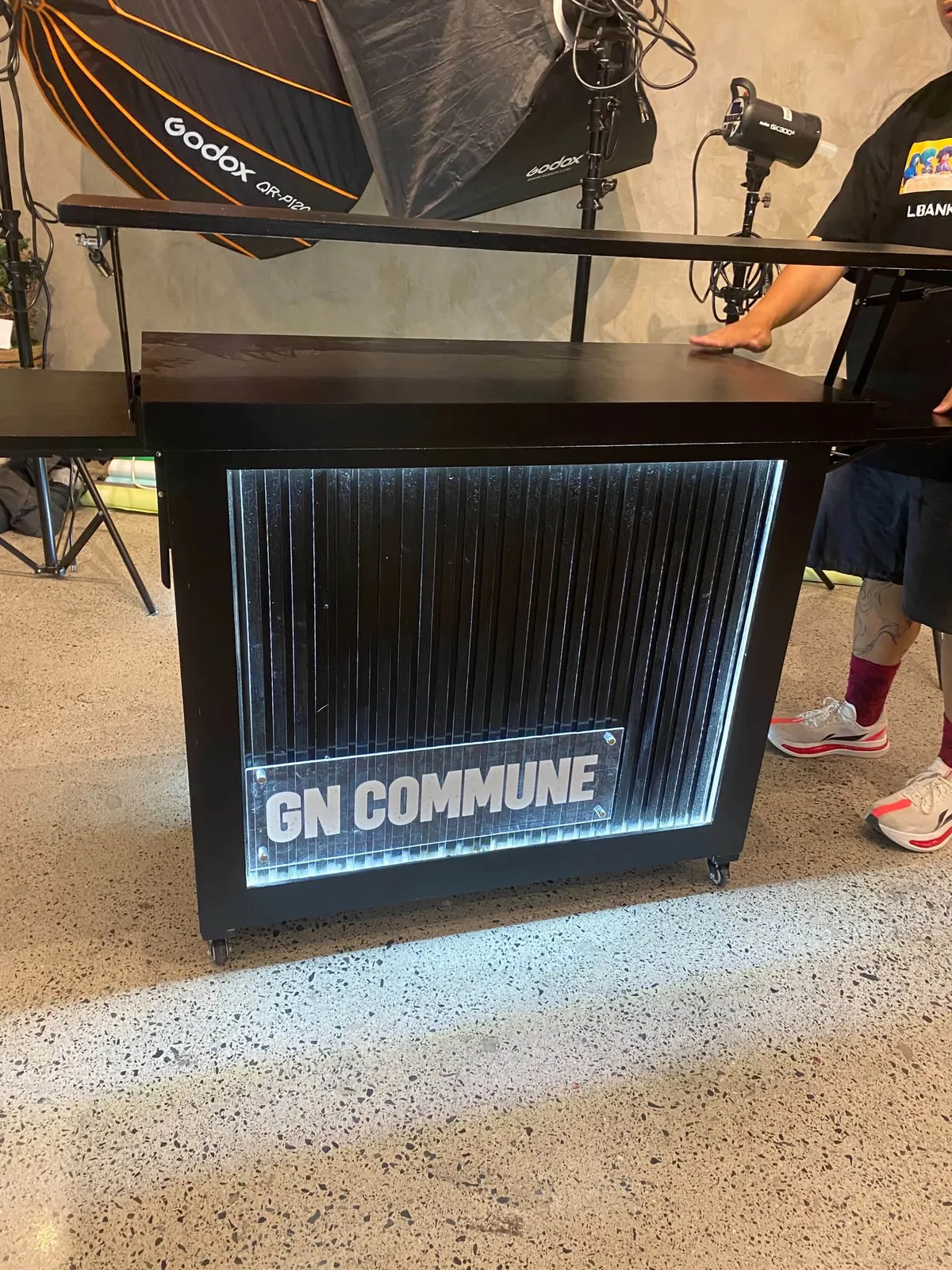 The front of the GN Commune cafe cart, a black cart on small wheels with a backlit ribbed glass panel and a clear GN COMMUNE sign, standing in a studio room with lighting stands behind it.