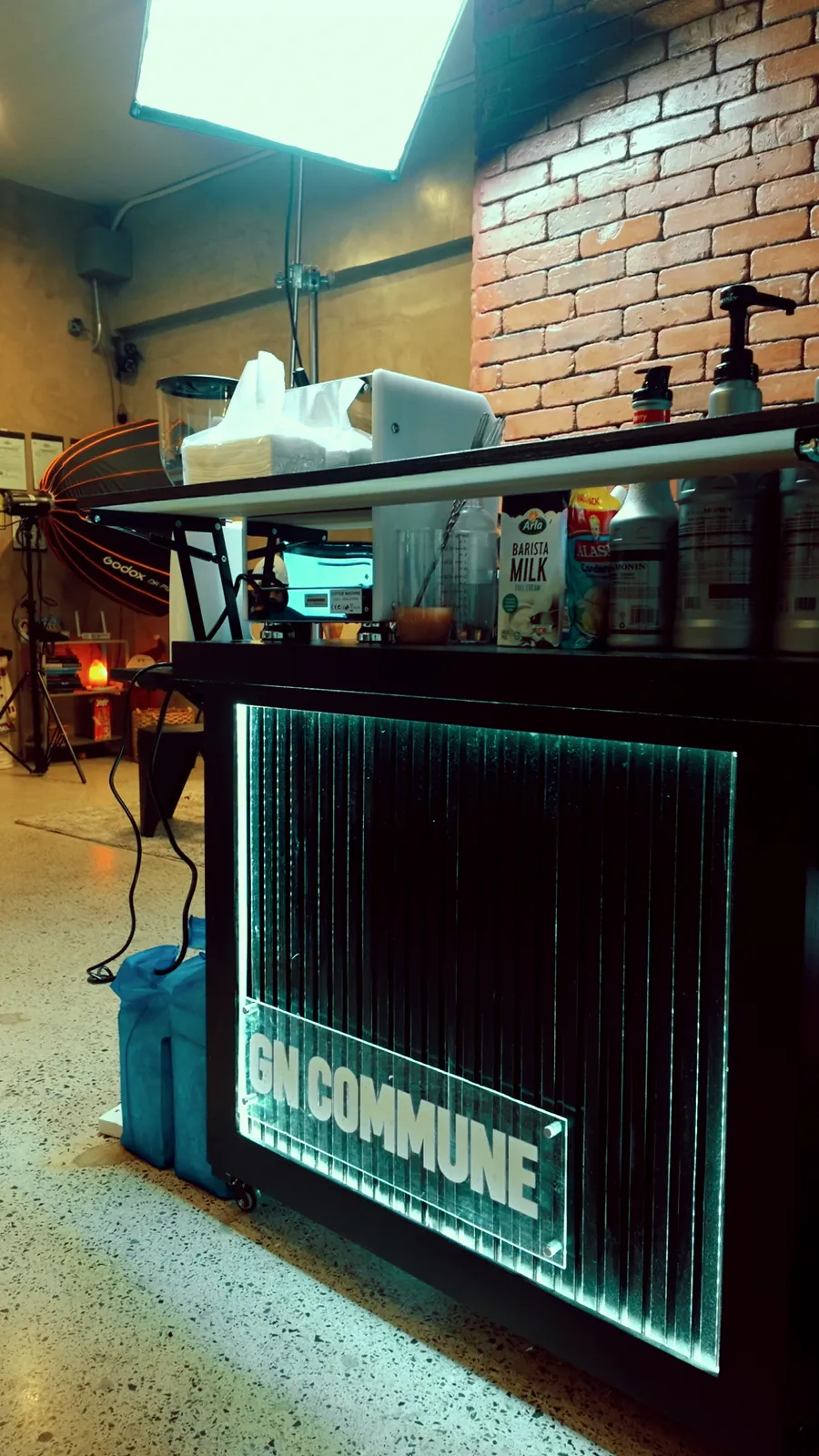 The GN Commune cart from the side: a black cart with a backlit ribbed panel and GN COMMUNE sign, syrups and milk on top, in front of a brick wall.