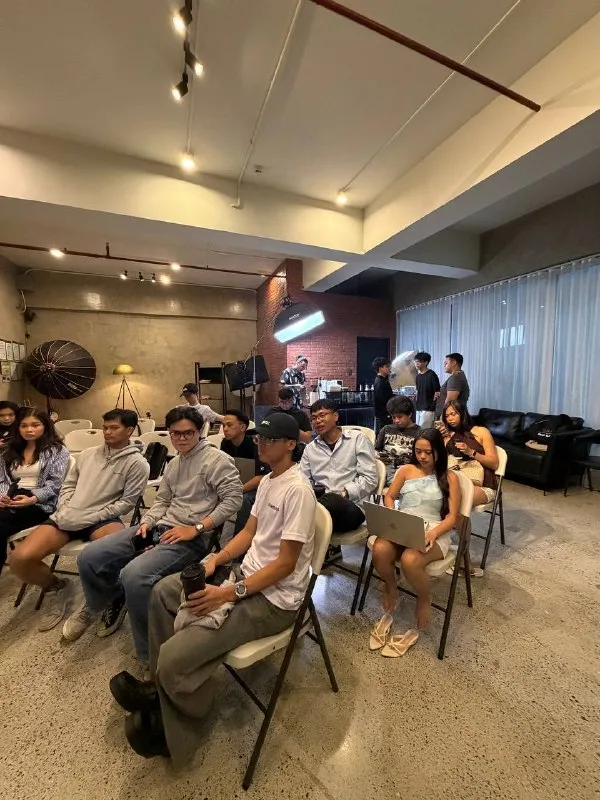 An audience seated in a brick and concrete room under studio lights, some of them holding coffee.