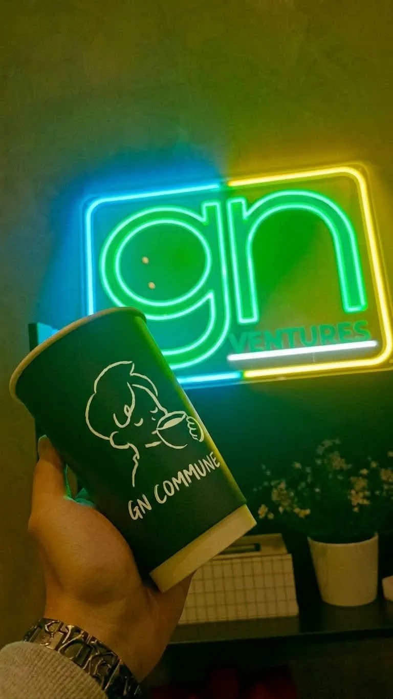A hand holding the GN Commune paper cup, a dark cup with a white line art doodle of a person sipping coffee, in front of a green, blue and yellow neon gn VENTURES sign.