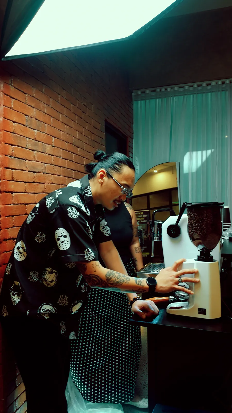 A barista in a black patterned shirt grinding beans at the cart beside a brick wall, a second barista working behind.