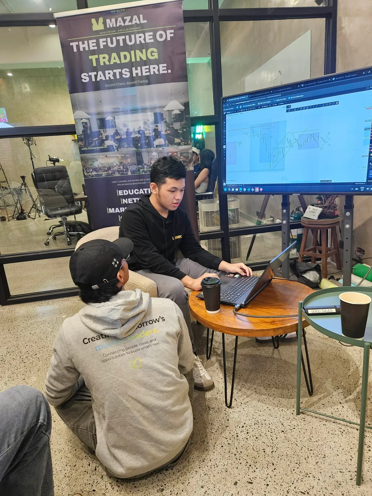 Two people at a laptop on a round wooden table with coffee cups, a monitor and a banner behind them.