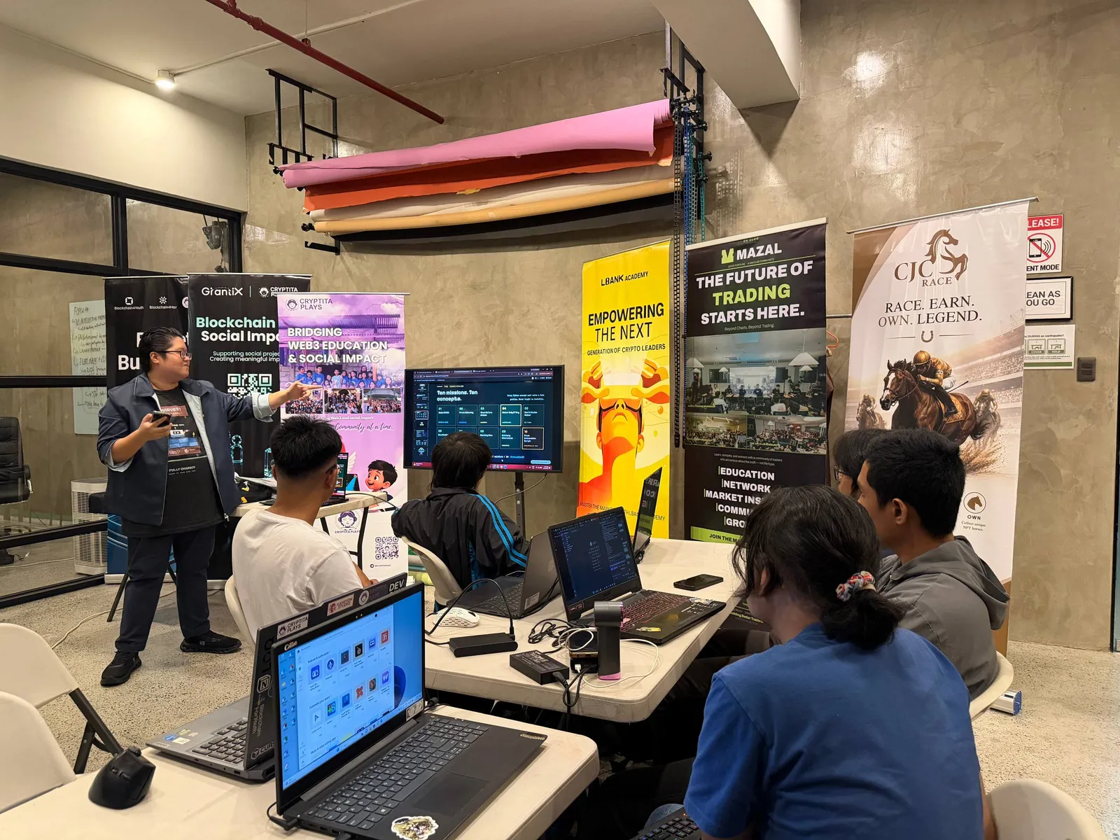A speaker at a screen while attendees sit at tables using laptops, with roll up banners behind them.