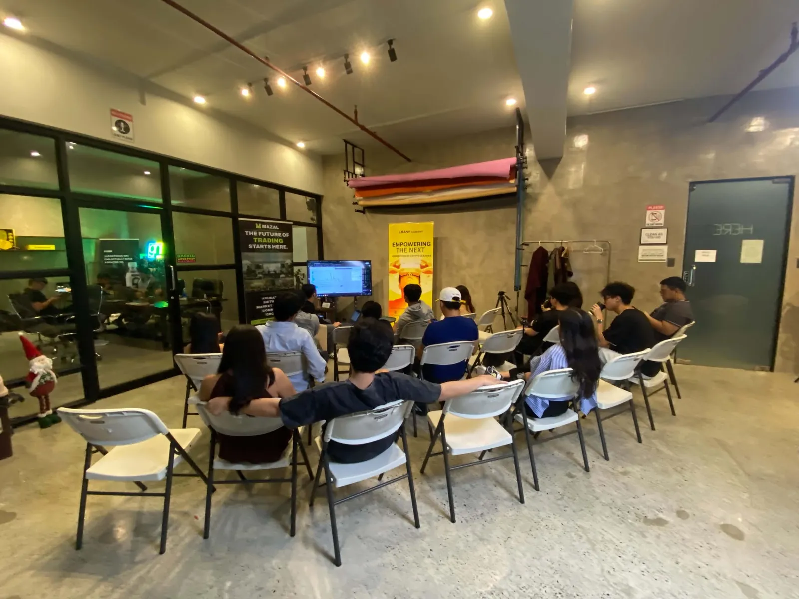 A seated audience on white folding chairs facing a screen in a loft style room with roll up banners and track lighting.