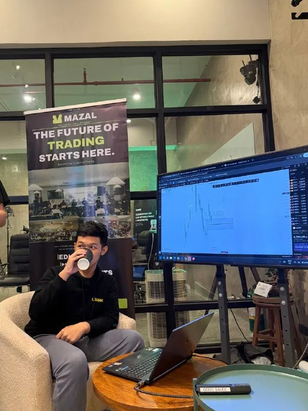 A person sipping from a cup at a table with a laptop and a big monitor.