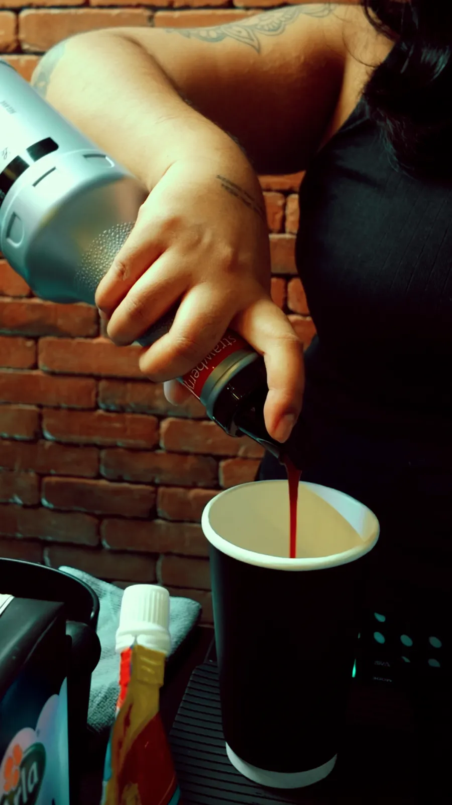 A barista pouring strawberry syrup into a black paper cup in front of a brick wall.